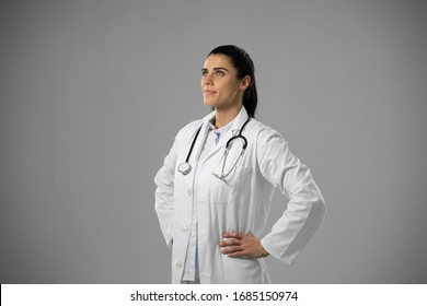 Side View Of A Happy Caucasian Female Doctor Wearing A Lab Coat With Stethoscope, Standing With Her Hands On Hips On A Grey Background.