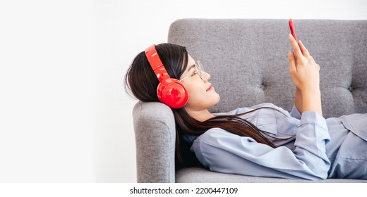 Side View Of Happy Cheerful Asian Woman Wearing Wireless Headphones Listening To Music From Smartphone Lay Down On Sofa.