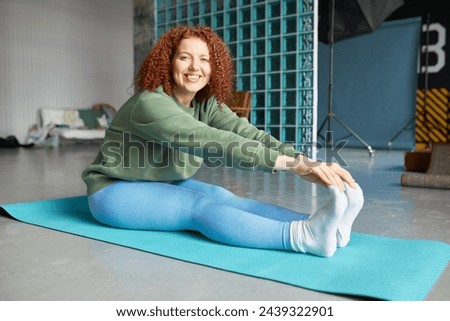 Side view of happy cheerful woman with red curly hair exercising on mat at gym, stretching legs and back side of hips reaching hands to feet and touching toes, looking at camera with smile