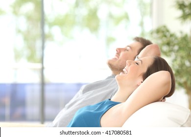 Side view of a happy couple breathing and resting lying in a couch at home with a window in the background