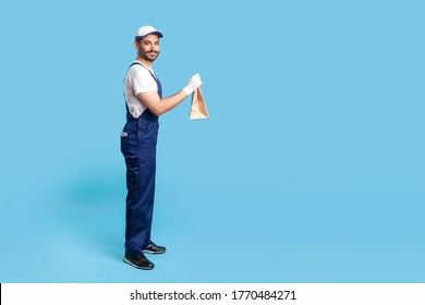 Side view, happy courier in blue uniform holding groceries bag and smiling to camera, carrying parcel with goods ordered online. Professional delivery service, food purchase. studio shot isolated