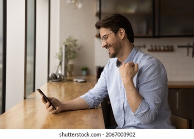 Side View Happy Emotional Young Caucasian Man Celebrating Online Lottery Gambling Auction Giveaway Win, Getting Victory Notification On Cellphone, Feeling Joyful Sitting At Table In Modern Kitchen.