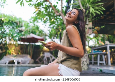Side view of happy ethnic female in eyeglasses sitting with head thrown back on edge of poolside while browsing cellphone in tropical resort on terrace with blurred background