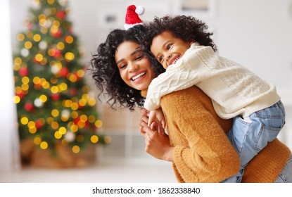 Side view of happy ethnic woman giving piggyback ride to boy and smiling while having fun at home during Christmas celebration