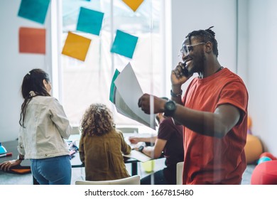 Side View Of Happy Excited African American Male With Documents In Hand Having Phone Conversation While Standing In Meeting Room With Colleagues Working Together In Background