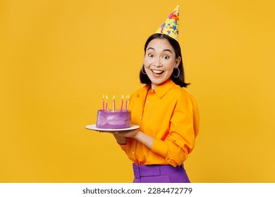 Side view happy fun smiling young woman wearing casual clothes cap hat celebrating holding purple cake with candles look camera isolated on plain yellow background. Birthday 8 14 holiday party concept