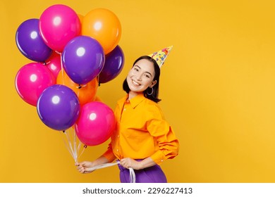 Side view happy fun smiling young woman wears casual clothes hat celebrating hold bunch of colorful air balloons look camera isolated on plain yellow background. Birthday 8 14 holiday party concept
