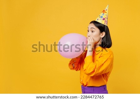 Side view happy fun young woman of Asian ethnicity wears casual clothes cap hat celebrating blowing pink balloon looking aside isolated on plain yellow background. Birthday 8 14 holiday party concept