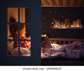 Side View Of Happy Girl Smiling And Reading Fairytale While Resting On Comfortable Bed Near Fairy Lights In Cozy Bedroom At Night At Home