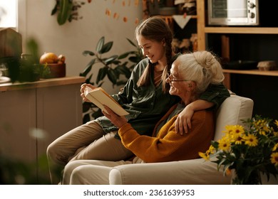 Side view of happy grandmother and granddaughter reading book of stories or novel at leisure while sitting in comfortable armchair - Powered by Shutterstock