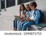 Side view of happy group of multiethnic school kids preteen boys and girls elementary middle pupils children sitting on the stairs and using digital devices laptop tablet smartphone outdoors