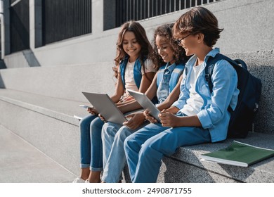 Side view of happy group of multiethnic school kids preteen boys and girls elementary middle pupils children sitting on the stairs and using digital devices laptop tablet smartphone outdoors - Powered by Shutterstock