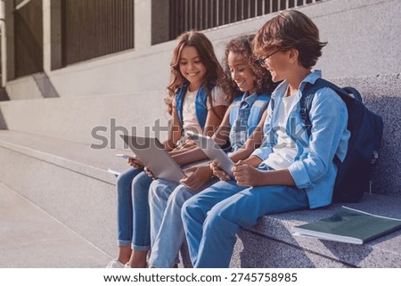 Side view of happy group of multiethnic school kids preteen boys and girls elementary middle pupils children sitting on the stairs and using digital devices laptop tablet smartphone outdoors