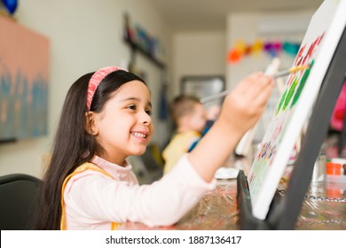 Side View Of A Happy Latin Girl Kid Painting A Colorful Landscape With A Brush On A Canvas During Her Art Class For Children