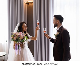 Side View Of Happy Loving Couple, Bride With Veil Wearing In Wedding Dress, Holding Bouquet Of Flowers, Standing With Elegant Groom In Suit While Holding Glass Of Drink Together Celebrating Engagement