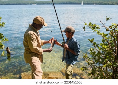 Side View Of Happy Mature Black Man In Activewear Showing Fish To His Grandson With Rod While Both Standing Against Waterside