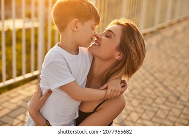 Side View Of Happy Mother Cuddling Face To Face With Adorable Boy And Touching Noses In Street In Summer Evening