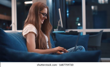 Side view of happy red hair young woman in white t-shirt sitting on blue coach browsing internet chatting with friends on modern laptop smile resting at modern apartment having fun evening relax - Powered by Shutterstock