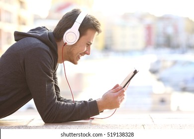 Side view of a happy teenager listening music or watching videos on line in a tablet lying on the floor with a port in the background - Powered by Shutterstock