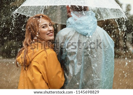 Side view of happy woman strolling with man. They are hiding under umbrella with joy. Smiling female is turning and looking back with content