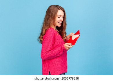 Side View Of Happy Woman Wearing Pink Pullover, Reading Letter Or Greeting Card, Holding Envelope, Smiling And Rejoicing Pleasant News. Indoor Studio Shot Isolated On Blue Background.