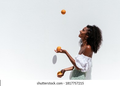 Side view of happy young African American female in summer outfit juggling with ripe oranges against white wall in sunny day