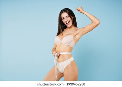 Side view of happy young brunette woman 20s in beige brassiere underwear standing posing measuring waist with measure tape doing winner gesture isolated on pastel blue background studio portrait