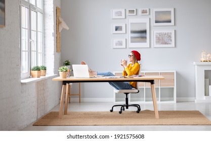 Side view of happy young woman enjoying hot drink holding legs on table and smiling while sitting on chair during break in work in creative workspace - Powered by Shutterstock