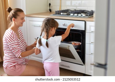 Side view of happy young woman and cute kid daughter wearing casual attires bake dessert in kitchen, teaching child making cupcakes, sitting on floor near oven stove, cooking at home.