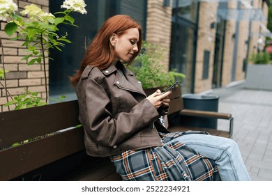 Side view of happy young woman in trendy leather jacket sitting on bench outdoors, happily browsing smartphone. Stylish redhead female using mobile phone on city bench, enjoying free time in city. - Powered by Shutterstock