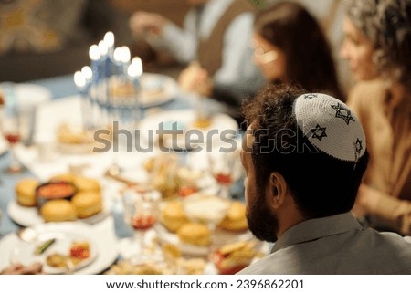 Side view of head of mature bearded man in yarmulke with stars of David sitting in front of camera by table served for Hanukkah dinner