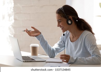 Side view head shot smiling mixed race lady freelancer wearing headset, communicating with client via video computer call. Millennial pleasant professional female tutor giving online language class.