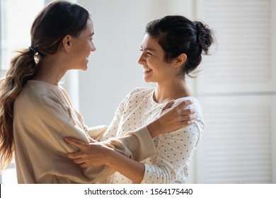 Side View Head Shot Two Mixed Race Girls Cuddling, Enjoying Communicating Together. Cheerful Indian Young Woman Talking With Best Friends, Discussing Life Events, Congratulating With Success.