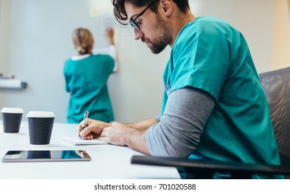 Side View Of Healthcare Worker Sitting In Hospital Boardroom Writing Notes With Female In Background.