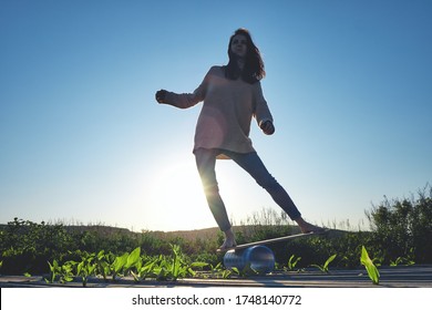 side view of healthy beautiful active woman keeping balance on the wooden balance board against the sky background at sunset summer day. Fitness and outdoor concept.