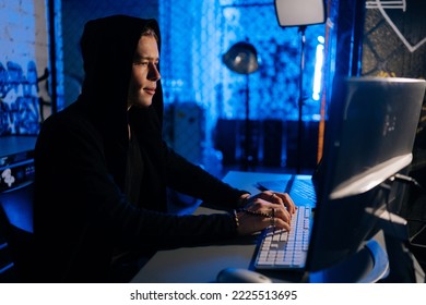 Side view of hidden hacker man wearing sweatshirt with hood engaged in hacking into security systems, sitting in dark room with blue neon lights. Concept of cyberattack, malware and cyber security. - Powered by Shutterstock