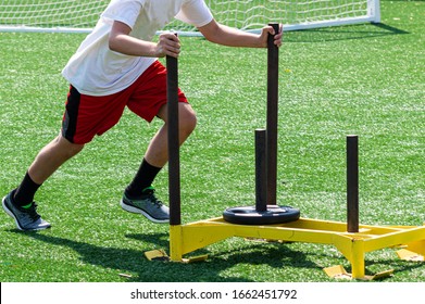 Side View Of A High School Boy Pushing A Yellow Sled With Weight On It For Resistance Training