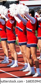 Side View Of High School Cheerleaders Holding Their Pom Poms Up Over Head While Cheering At Their Homecoming Football Game.