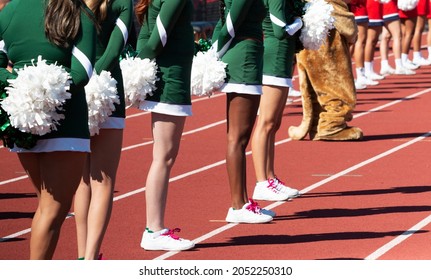 Side View Of High School Cheerleaders From Both Teams Standing On The Track Infront Of The Stands During A Football Game