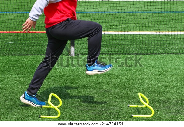 Side view of a high school track runner\
standining in the track A position over a small yellow hurdle doing\
sports training drills.