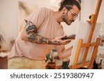 Side view of a hipster artist standing at creative art studio with palette in hands and creating an unique artwork on easel. Talented tattooed male painter painting on canvas at creative atelier alone