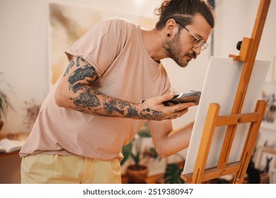 Side view of a hipster artist standing at creative art studio with palette in hands and creating an unique artwork on easel. Talented tattooed male painter painting on canvas at creative atelier alone - Powered by Shutterstock