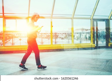 Side View Of Hipster Male Using App On Mobile Phone And Sending Message While Walking In Back Lit On Covered Pedestrian Bridge In City