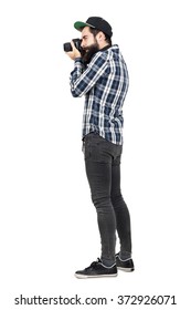 Side View Of Hipster In Plaid Shirt And Baseball Cap Taking Photo With Dslr Camera. Full Body Length Portrait Isolated Over White Studio Background.