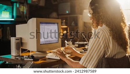 Side View Of Hispanic Female Engineering Student Writing Academic Thesis On Old Desktop Computer In Retro Garage. Future Engineer Focused On Innovative Project For University Of Technology In Nineties