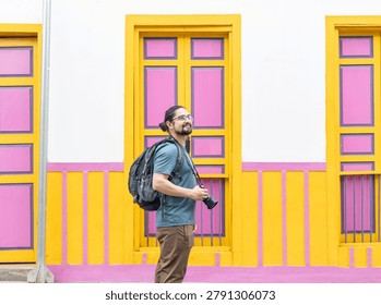 Side view of a Hispanic man with backpack holding a camera in front of traditional pink and yellow windows and doors.