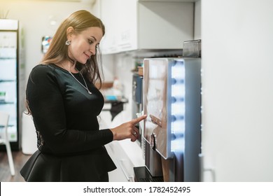 Side view of a hispanic woman with long hair choosing what type of coffee drink to prepare using a coffee machine maker while standing indoors of a cozy office kitchen during a break