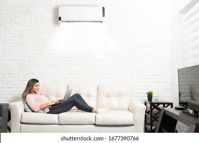 Side view of Hispanic woman using laptop while reclining on sofa below air conditioner at home - Shutterstock ID 1677390766