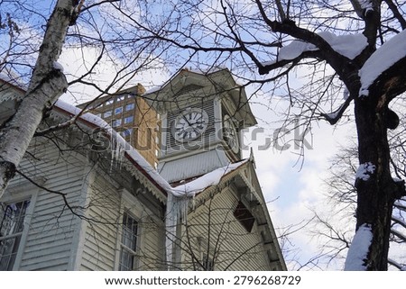 A side view of the historic Sapporo Clock Tower partially framed by winter tree branches and surrounding city buildings.