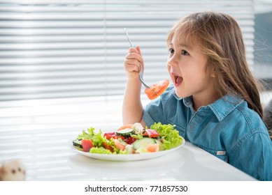 Side View Of Hungry Asian Girl Eating Piece Of Tomato With Appetite. She Is Sitting At Table In Kitchen